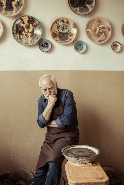 Senior potter in schort denken en permanent tegen muur met hangende aardewerk goederen - Foto, afbeelding