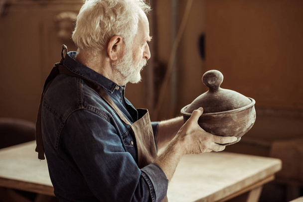 Старший гончар в фартуке рассматривает керамическую миску в мастерской
 - Фото, изображение