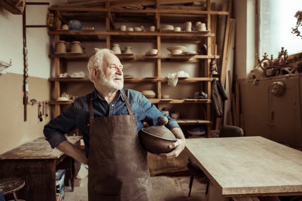 Vista frontal do oleiro sênior orgulhoso em pé avental com tigela de cerâmica na oficina
 - Foto, Imagem