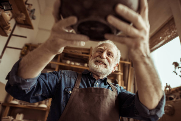 Alfarero senior en delantal examinando tazón de cerámica en el taller
 - Foto, Imagen