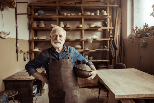 Vista frontal del orgulloso alfarero senior en delantal de pie con cuenco de cerámica en el taller
 - Foto, Imagen