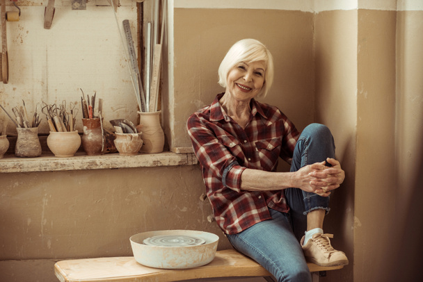 Feliz mujer mayor sentada en el banco cerca de la pared en el taller
 - Foto, imagen