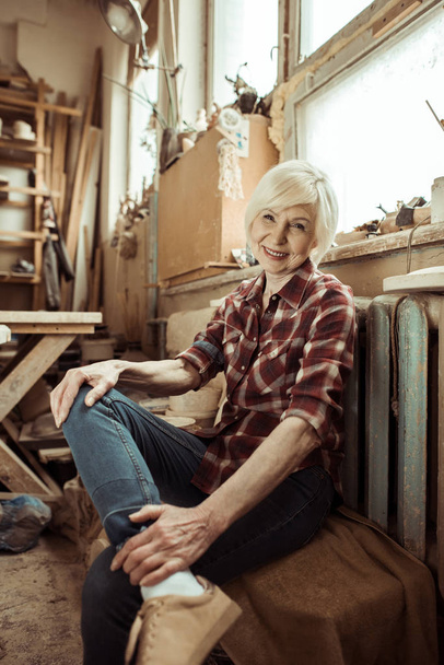 Feliz mujer mayor sentada en el banco cerca de la ventana en el taller
 - Foto, Imagen