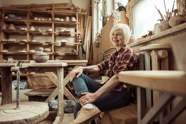 Happy senior woman sitting on bench near window at workshop - Zdjęcie, obraz