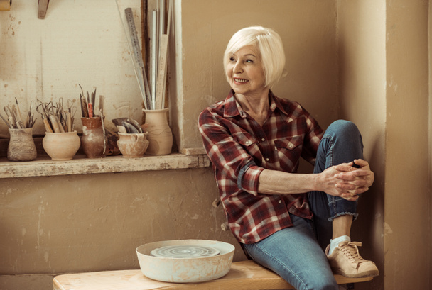 Heureuse femme âgée assise sur un banc près du mur à l'atelier
 - Photo, image