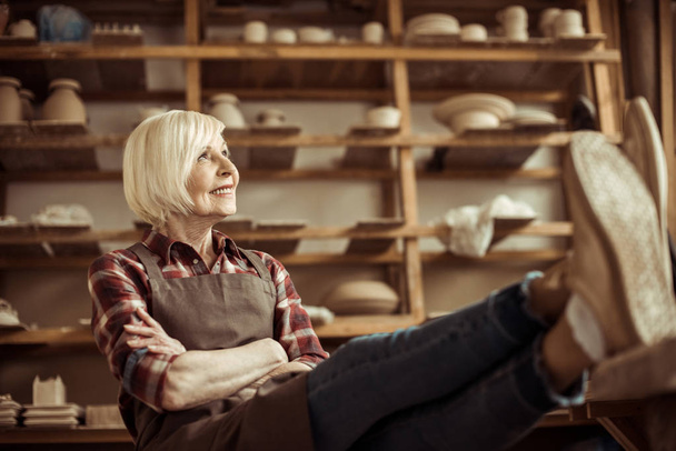 Vista frontal de la mujer mayor sentada en silla con patas en la mesa contra los estantes con productos de cerámica
 - Foto, Imagen