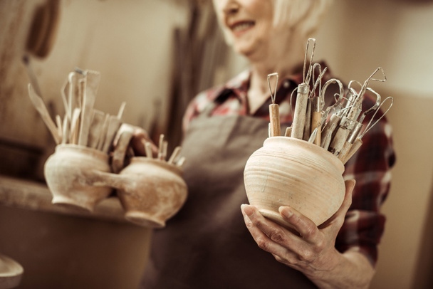Mujer mayor sosteniendo cuencos con herramientas de cerámica en el taller
 - Foto, Imagen