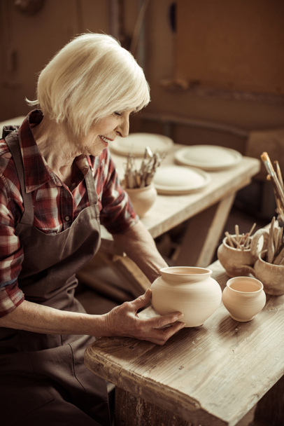 Female potter sitting at table and examining ceramic bowl at workshop - Photo, Image