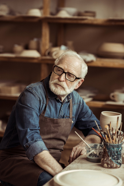 Senior potter in apron sitting at table and daydreaming at workshop - Foto, Imagem