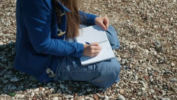 Una mujer está sentada en una playa pedregosa y escribiendo un bolígrafo en el cuaderno
. - Imágenes, Vídeo