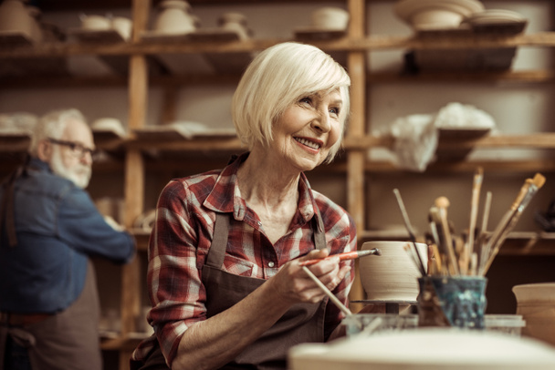 Mujer pintando olla de barro con alfarero senior en el taller
 - Foto, Imagen