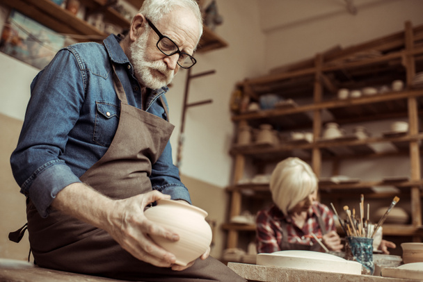 Senior-Töpfer in Schürze und Brille begutachtet Keramikschale mit Frau im Hintergrund - Foto, Bild