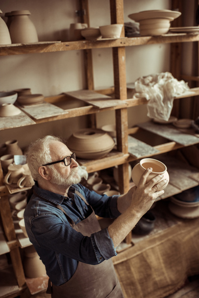 Alfarero senior en delantal y anteojos examinando cuenco de cerámica en el taller
 - Foto, Imagen
