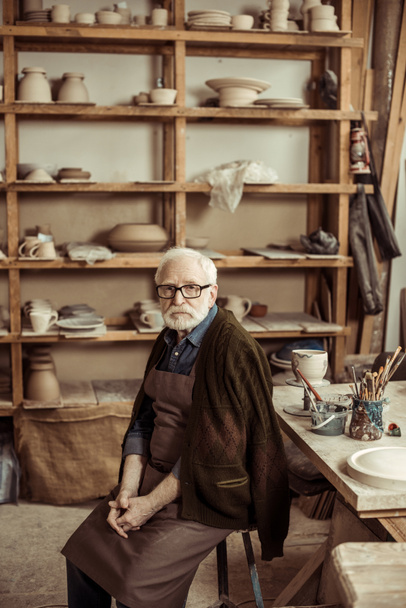 Senior potter in apron sitting at table and looking at camera at manufacturing - Photo, Image