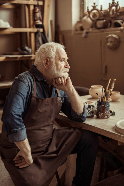 Senior potter in schort aan tafel zit en dagdromen in productie - Foto, afbeelding