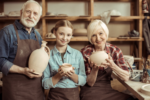 Nipote e nonni in piedi e in possesso di vaso di argilla e ciotole contro parete con ceramiche
 - Foto, immagini