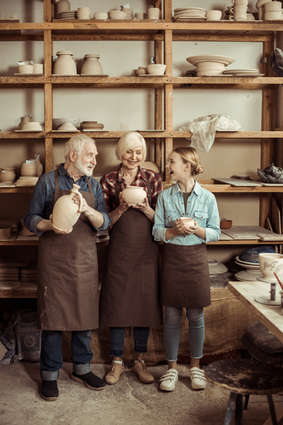 Granddaughter and grandparents standing and holding clay vase and bowls against wall with pottery goods - Foto, Imagem