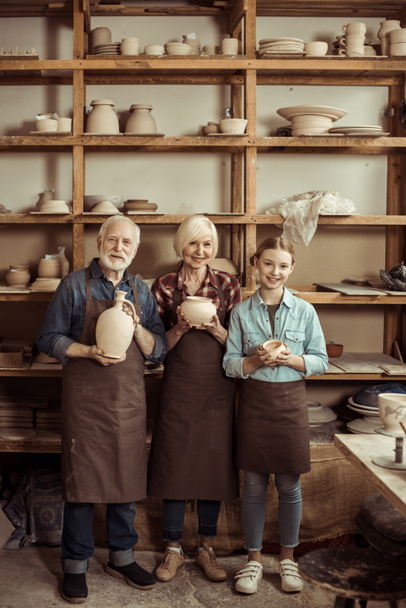 Granddaughter and grandparents standing and holding clay vase and bowls against wall with pottery goods - Photo, Image