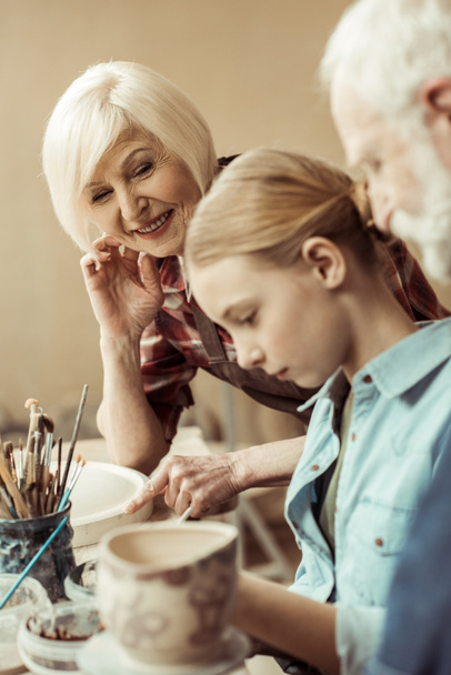 Vue latérale de fille peinture pot d'argile et grands-parents aider à l'atelier
 - Photo, image