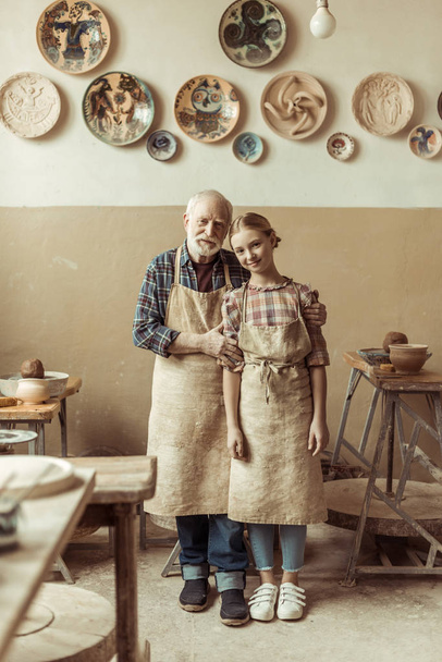 Front view of senior potter with his granddaughter in aprons standing at workshop - Photo, Image