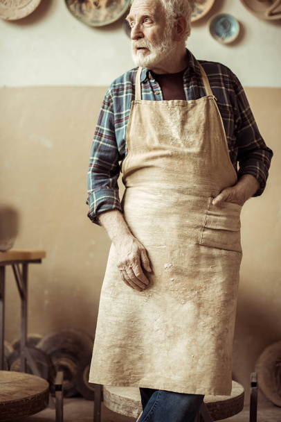 Front view of senior potter in apron standing at workshop - Photo, Image