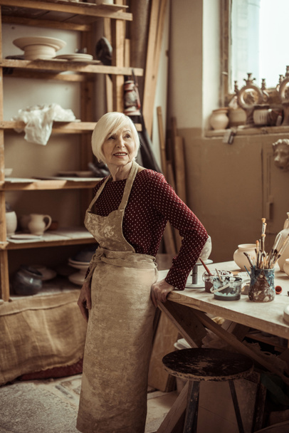 Front view of senior female potter in apron standing at workshop - Foto, Imagem