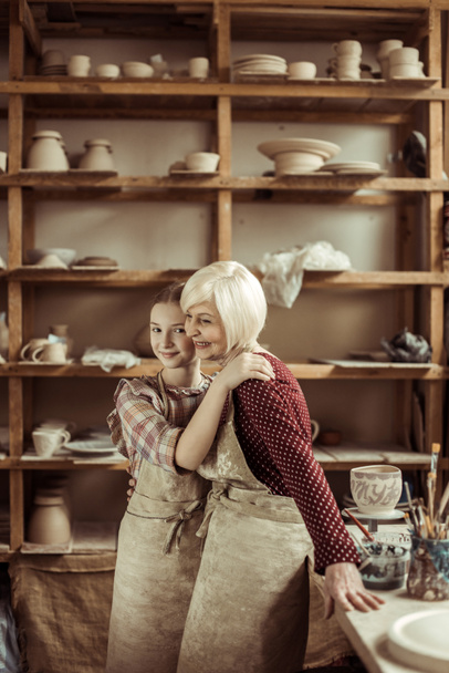 Внучка обнимает бабушку, пока они стоят в фартуках в мастерской
 - Фото, изображение