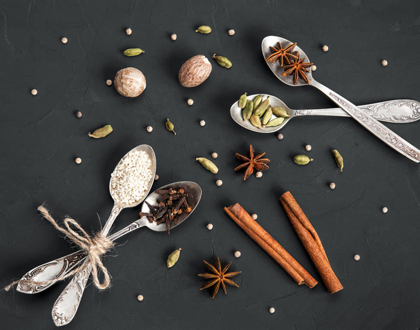 Various spices in metal vintage spoons and cinnamon sticks on dark rustic background. Cardamom, white pepper, nutmeg, anise, clove, sesame seeds.  - Photo, Image