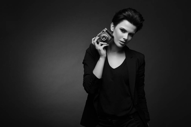 Young serious beautiful woman with short dark hair wearing black retro blazer posing in dark studio, holding old photo camera in her hands, in black and white - Fotografie, Obrázek