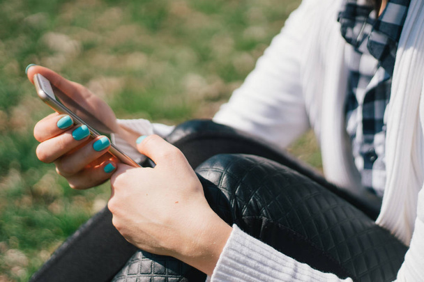 Beautiful young European girl sits on the grass in the park and uses a smartphone, concepts of using gadgets in a natural environment in the fresh air - Foto, afbeelding
