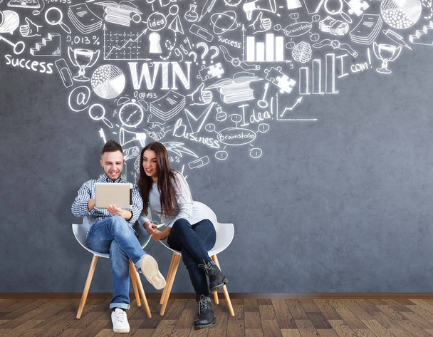 Young man and woman sitting on chairs and using laptop together in interior with business sketch on concrete wall. Teamwork concept - Photo, Image