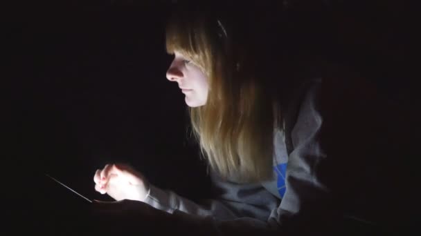 Young handsome blonde girl using her tablet laying on the bed in her lights off house - Imágenes, Vídeo