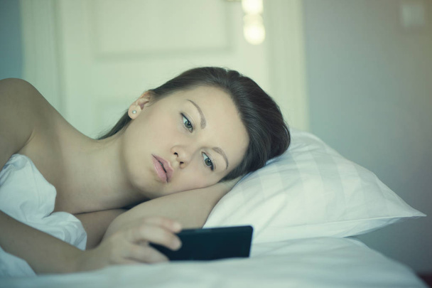 A beautiful girl lies in bed and can not fall asleep and reads news on the smartphone. Insomnia. Psychology. Phobias - Фото, зображення