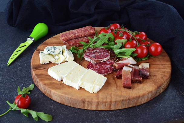 Olive wood cutting board with cherry tomatoes, arugula and variety of sliced cheeses and sausages. Snack concept. - Фото, изображение