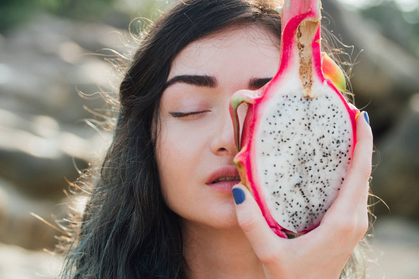 Girl is holding a dragon fruit next to her face. A pitaya fruit holds in hand on the beach. Tropical Dragon Fruit cut in half. - Фото, зображення