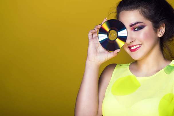 Portrait of young pretty girl with colourful artistic make-up and updo hair holding a CD in front of her eye and smiling - Photo, Image