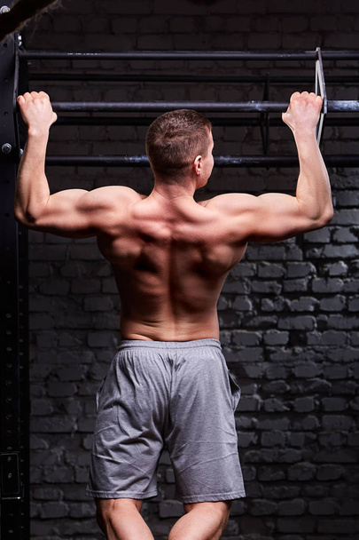 Rear view photo of muscular male doing exercises on horizontal bar against brick wall at the cross fit gym. - Photo, image