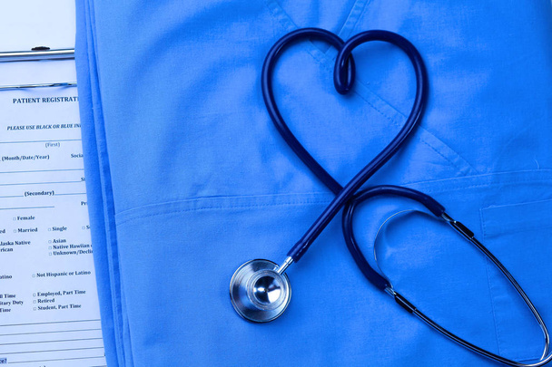 Medical stethoscope twisted in heart shape lying on patient medical history list and blue doctor uniform closeup - Photo, Image