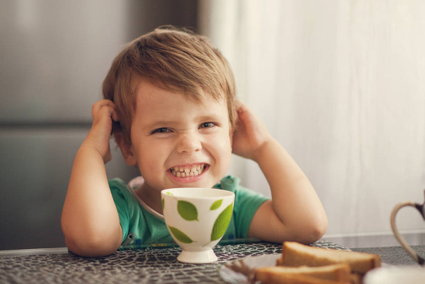 Cheerful boy drinks milk, eats toast for breakfast - Фото, изображение