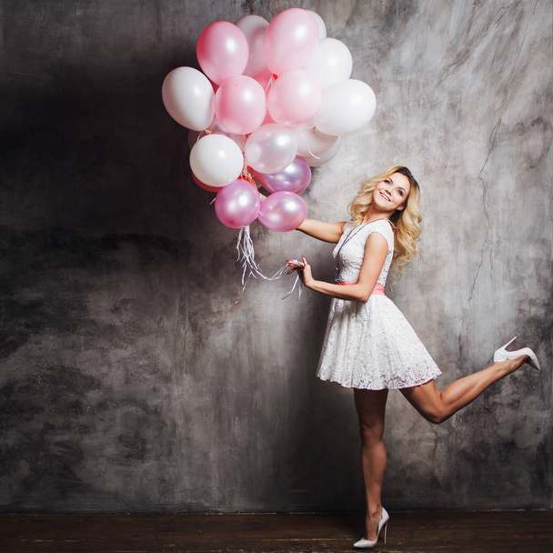 Charming young blonde in a white dress with pink sash, holding a large bundle of balloons at the party. - Foto, imagen