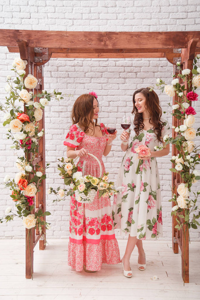 Duas meninas bonitas vestidas em vestidos de verão posando perto de um arco de flor com copos de vinho tinto nas mãos
 - Foto, Imagem