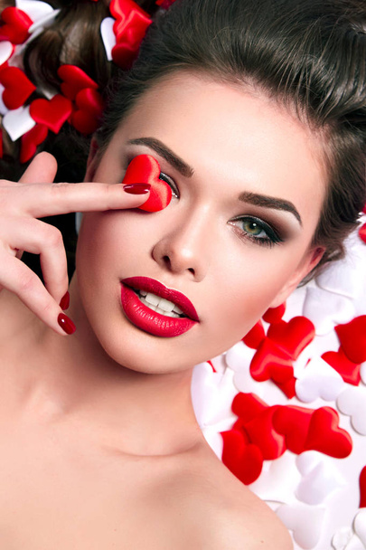 A beautiful young girl lies in small hearts. Smooth wavy hair, green eyes, red chubby lips. Celebration, advertising cosmetics, spa, romance. - Valokuva, kuva