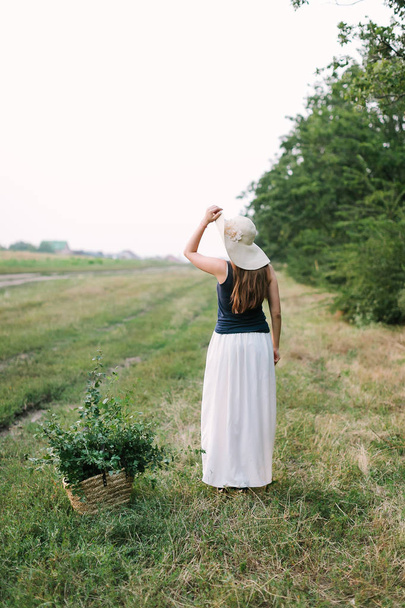 people, holidays and nature concept - beautiful woman in fancy clothes and elegant hat posing with back to us against rural fields and trees, on ground is basket with green branches for decoration. - Fotoğraf, Görsel