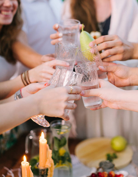 picnic, people, holiday concept - happy young people clink glasses near table with flagon of lemonade and candle, one glass with whole pear instead of lemonade, flagon with lemons, limes, mint leaves. - Photo, Image