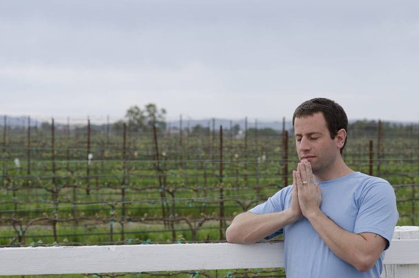 Mann betet angelehnt an einen weißen Zaun in einem Weinberg. - Foto, Bild