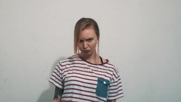 Chica rubia gruñona en camisa a rayas con tatuajes enfurruñados delante de la pared blanca
 - Metraje, vídeo