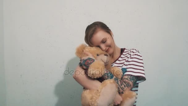 Roztomilá blondýnka v pruhované tričko s tetováním, objímání s hračka medvídek - Záběry, video