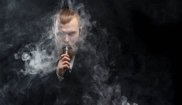 Vaping man holding a mod. A cloud of vapor. Black background. Studio shooting. - Фото, изображение