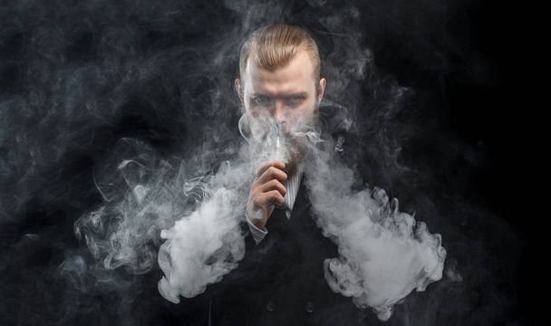 Vaping man holding a mod. A cloud of vapor. Black background. Studio shooting. - 写真・画像