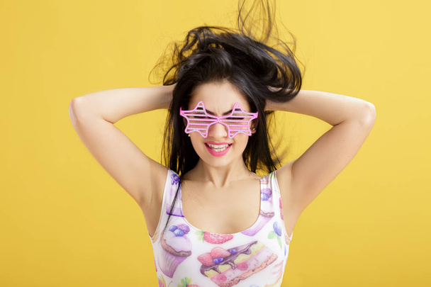 attractive young brunette woman in pink skirt and tank top on yellow background. funny girl with pink sunglasses - 写真・画像